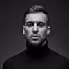 Black and white portrait of a man with short hair and a beard wearing a black turtleneck, against a dark background.